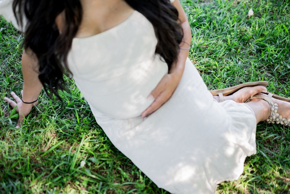 A pregnant woman in a white dress sits on green grass, cradling her baby bump with one hand. Family/Maternity Photography.