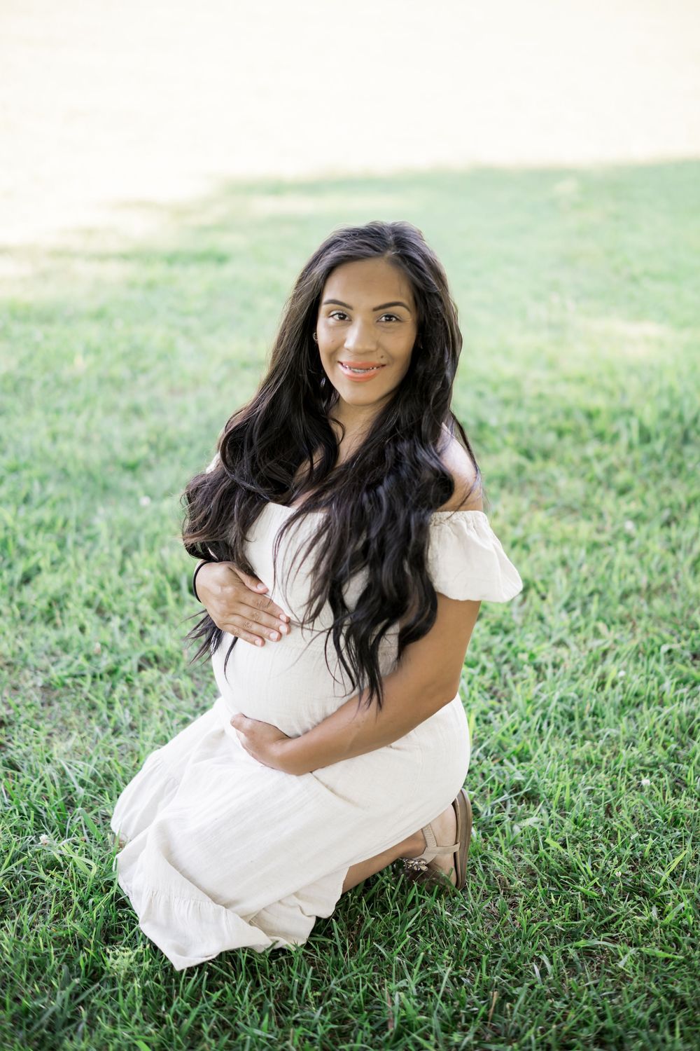 Pregnant woman in white off-shoulder dress kneeling on grass, cradling baby bump, long dark hair, smiling. Family/Maternity Photography.