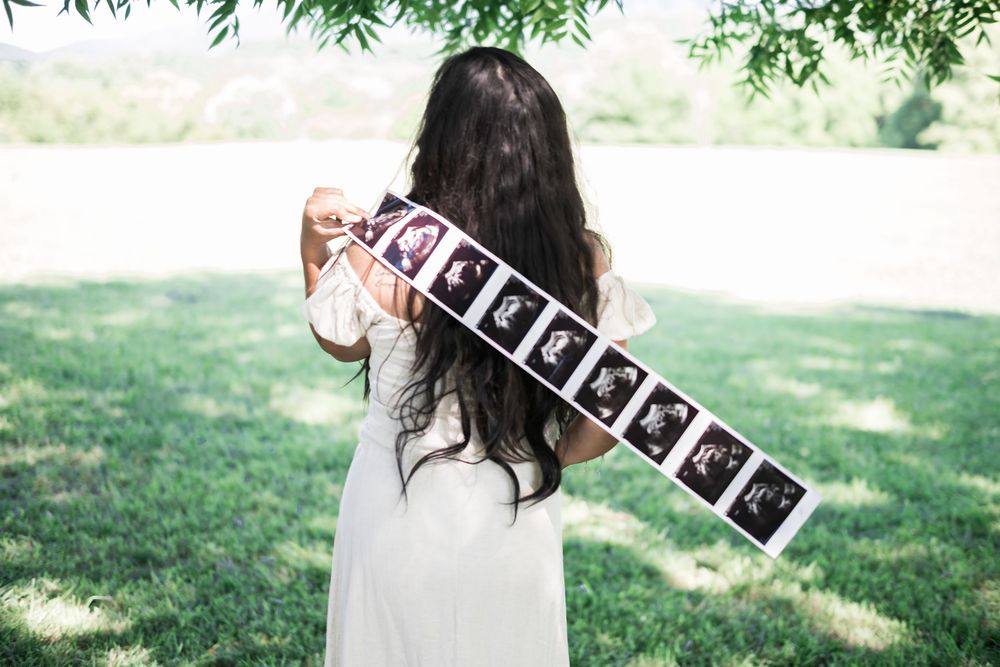 Pregnant woman in white dress holding ultrasound photo strip outdoors in green park setting. Family/Maternity Photography.