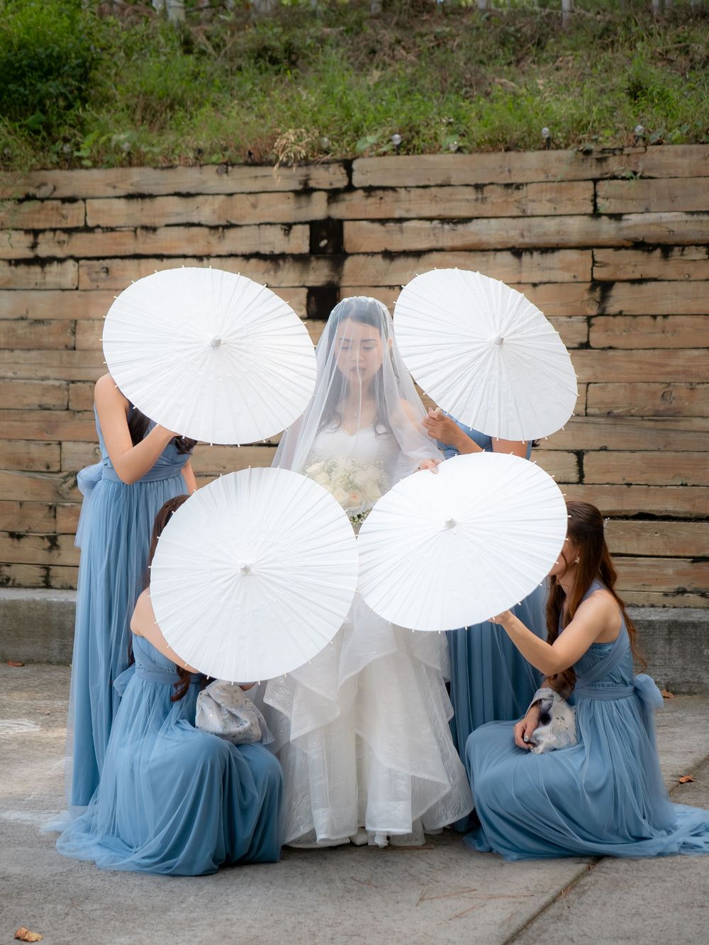 Wedding party in blue dresses holding white parasols while seated against rustic brick wall.