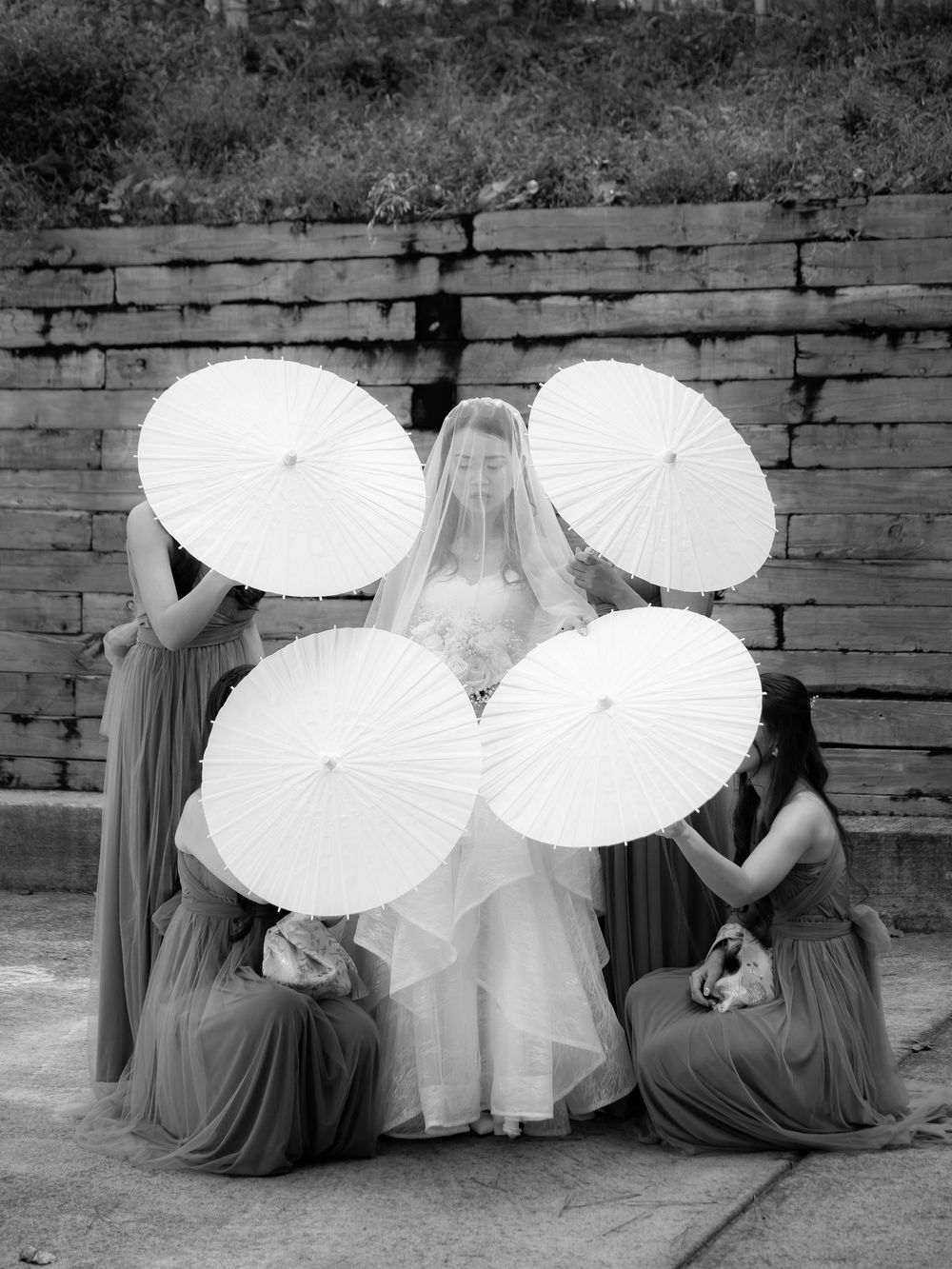 Black and white photo of wedding party seated with white parasols against brick wall.