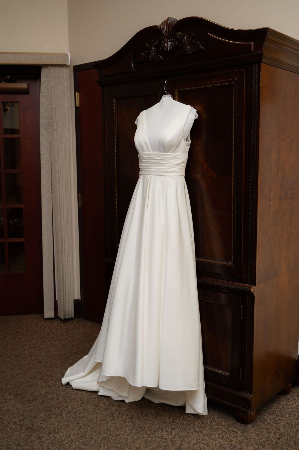 White wedding dress hanging on display against dark wooden paneling in a church dressing room.