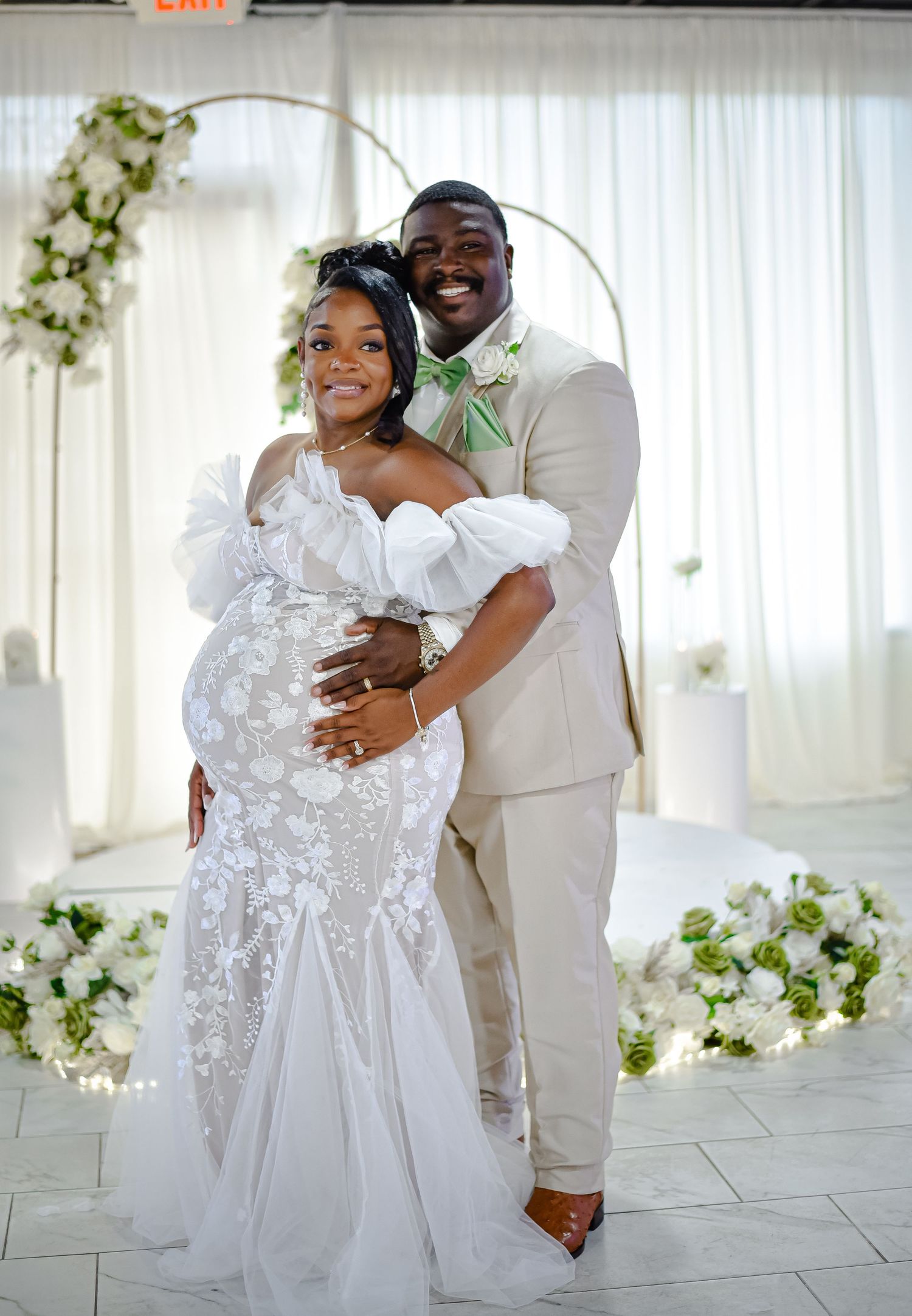 Happy couple embraces and laughs in elegant white wedding attire against floral backdrop.
