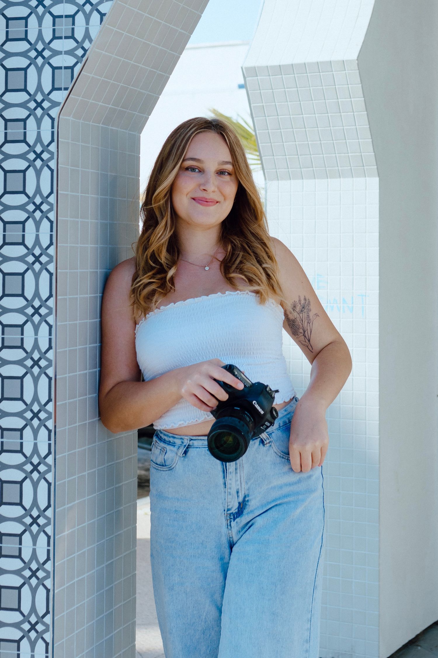 Photographer in white top and jeans holding camera against geometric tiled wall.