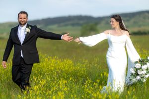 Two newlyweds spin joyfully in a field of yellow wildflowers on their wedding day with rolling hills in the background.