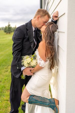 Romantic newlyweds share tender kisses against a white wall backdrop in the countryside.