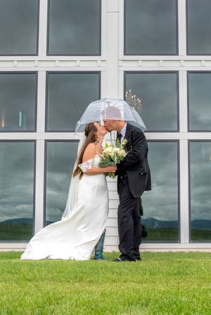 A bride and groom share romantic kisses under an umbrella against a stormy sky and modern building backdrop.