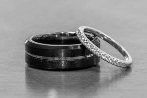 Black tungsten wedding band and delicate diamond eternity ring displayed together on a gray surface in black and white.