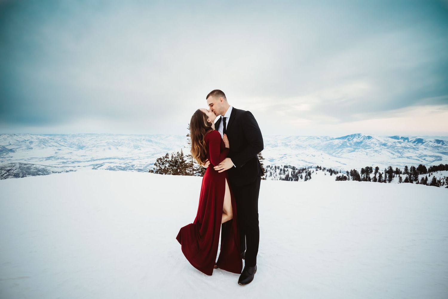 Woman in maroon dress with her husband in a black suit posing on a snow covered mountain in Utah