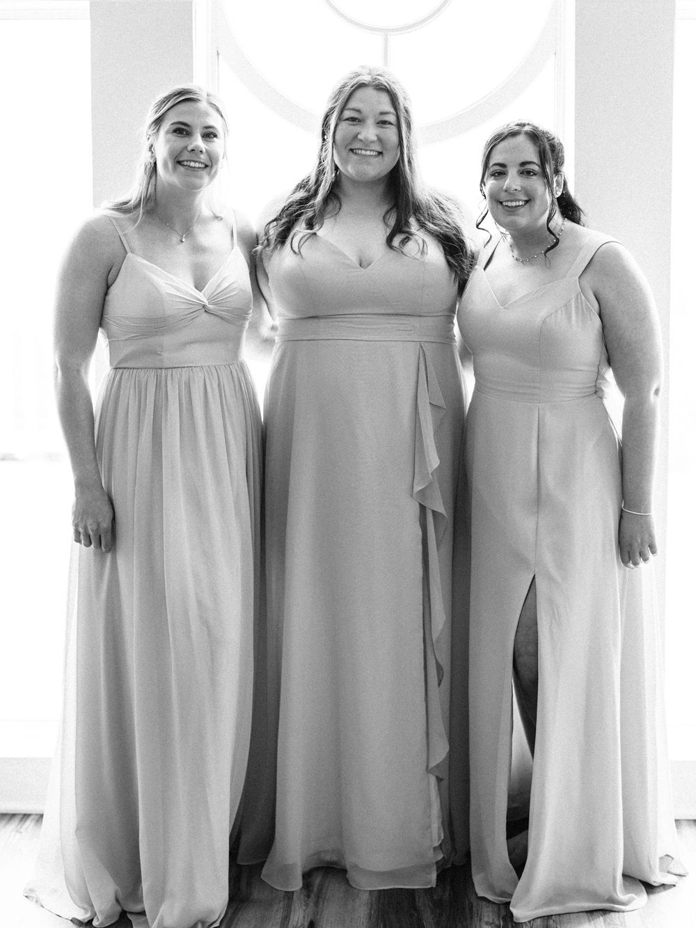 Three bridesmaids in light colored floor-length dresses pose together.