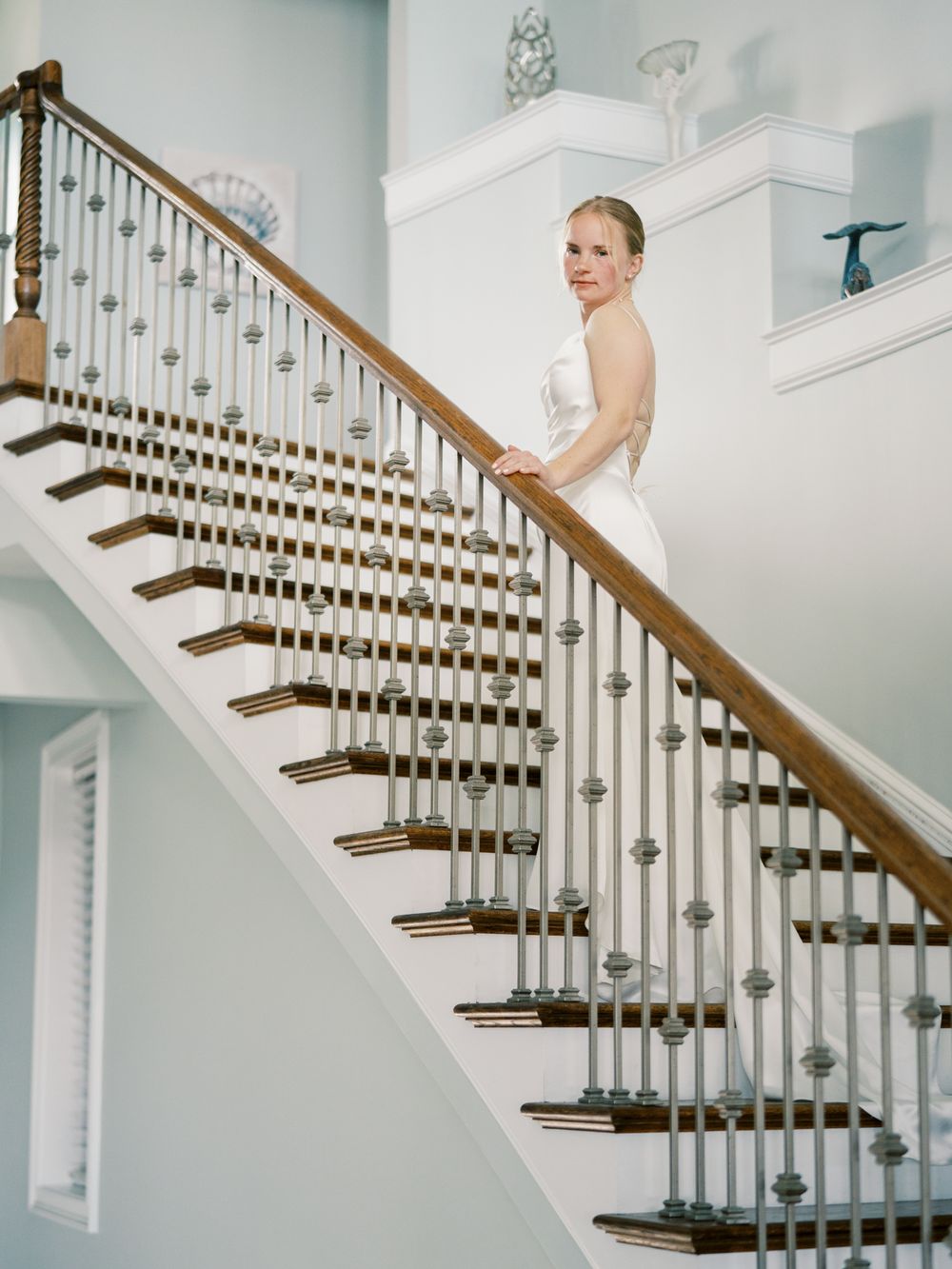 Bride in white dress stands on elegant wooden staircase with white spindles.