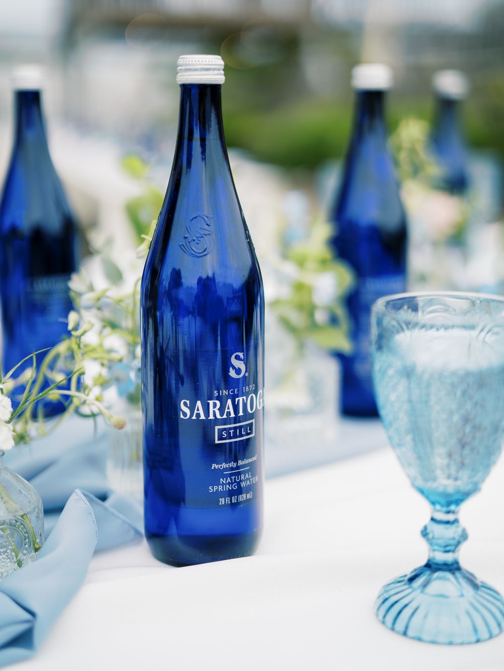 Blue Saratoga water bottle and vintage blue goblet on white tablecloth with greenery.