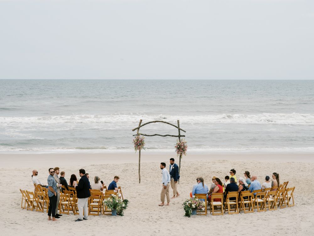 Beach wedding ceremony setup with wooden chairs and simple driftwood arch on sandy shoreline.