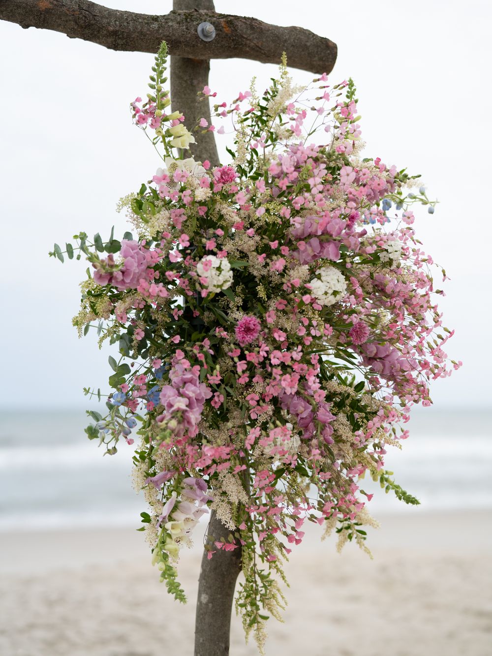 Lush pink and white floral arrangement mounted on driftwood arch at beach ceremony.