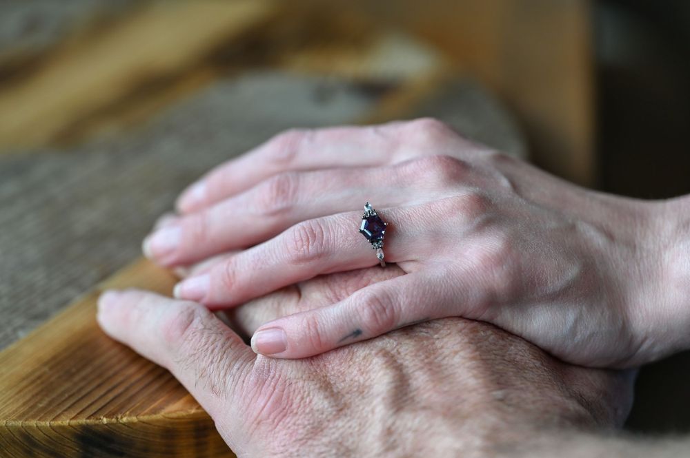Close-up of two hands resting together, one adorned with a sapphire and diamond engagement ring on a wooden surface.