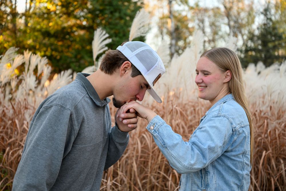 A young man in a white cap kisses the hand of a smiling woman in a denim jacket among golden pampas grass in an autumn field.