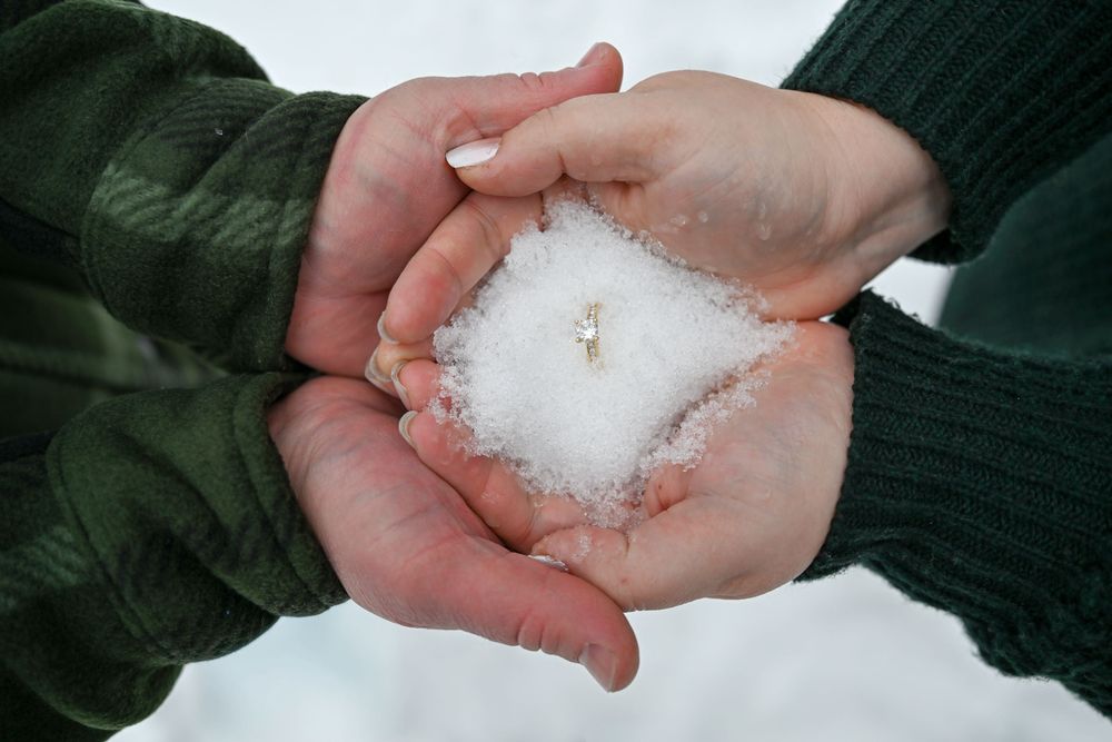 Couple cupping hands together holding snow with diamond engagement ring on top.