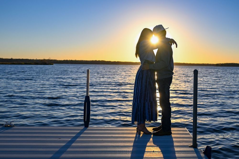 Silhouetted couple in a romantic kiss on a wooden lake dock at sunset, with the sun glowing between them over calm blue water.