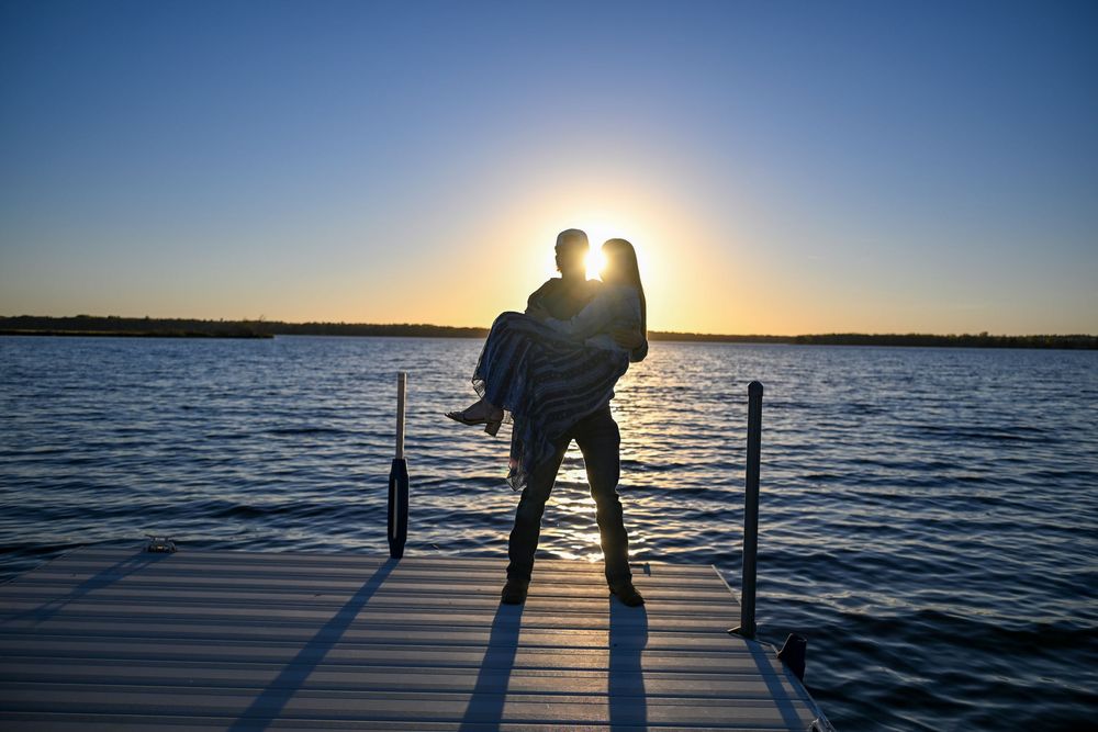 Silhouette of a man carrying a woman in his arms on a wooden dock at sunset, with calm lake water and a glowing sun on the horizon.