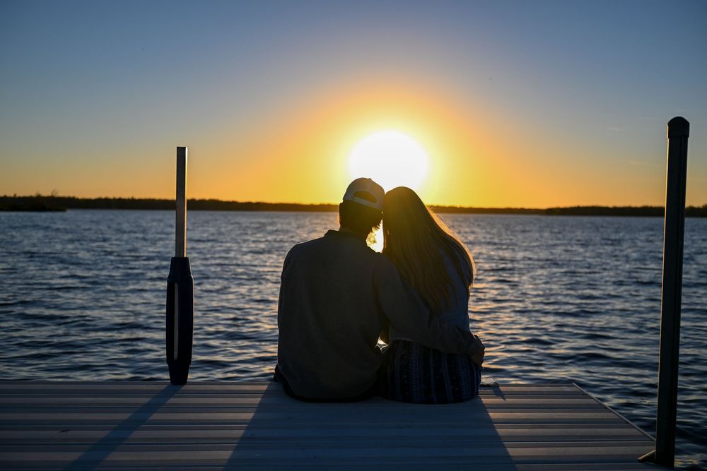 Silhouetted couple sitting close together on a wooden dock watching a golden sunset over calm lake water.