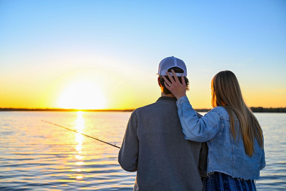 Couple stands at the water's edge fishing during a golden sunset, with her hand resting on his shoulder over a calm lake.