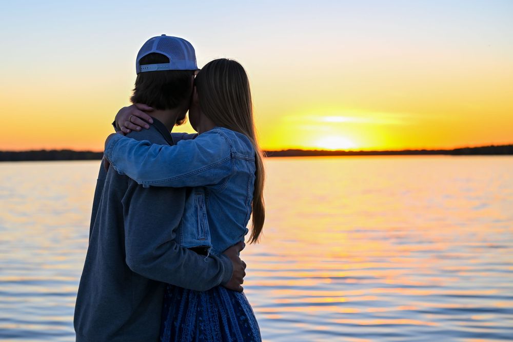 Couple embracing at the water's edge while watching a vibrant orange sunset over a calm lake.