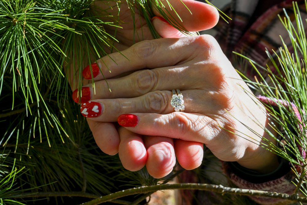 Close-up of hands showing a sparkling diamond engagement ring with red manicured nails among pine branches.