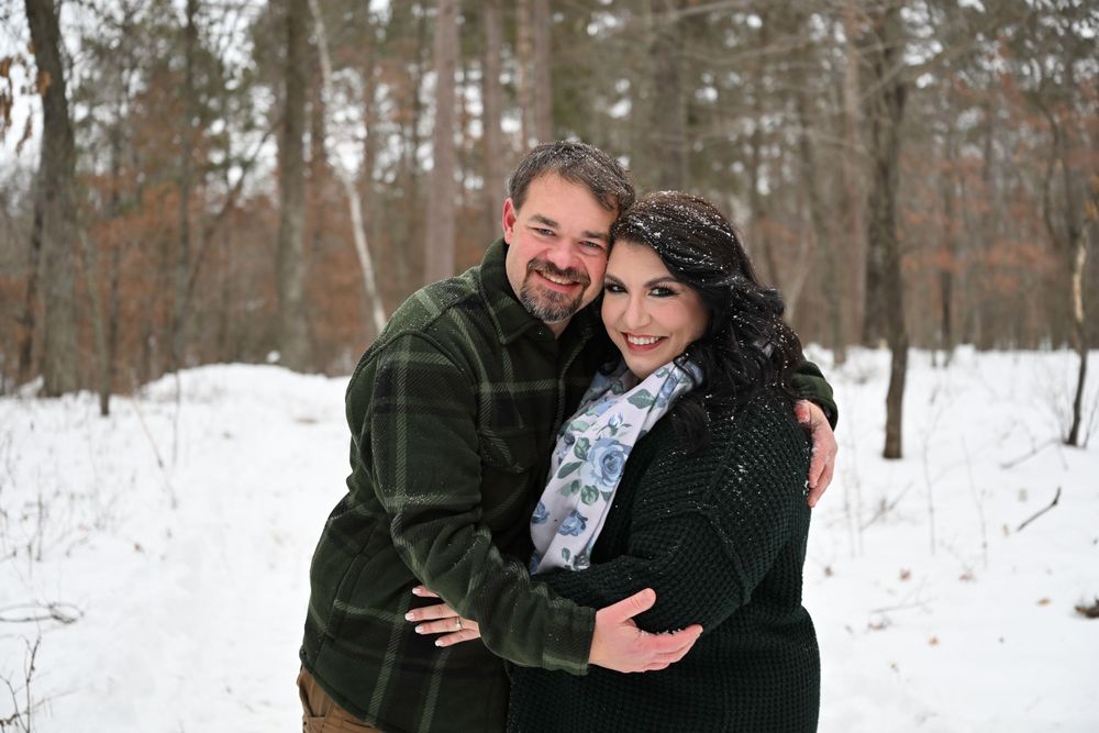 Couple embracing and smiling together in snowy winter forest during engagement photos.