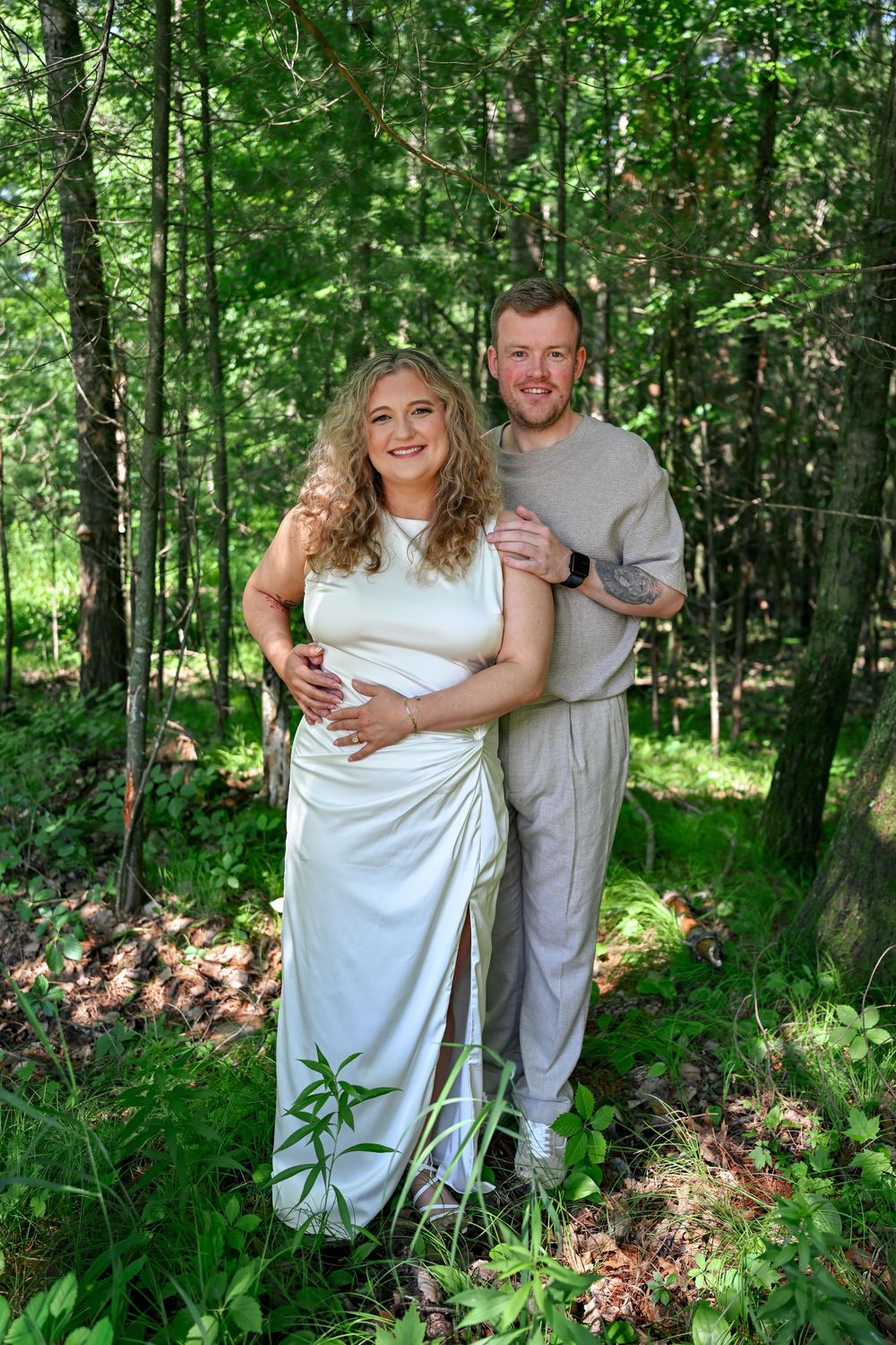 A couple poses in a lush green forest, the woman in a white wedding gown and the man in a gray outfit embracing her.