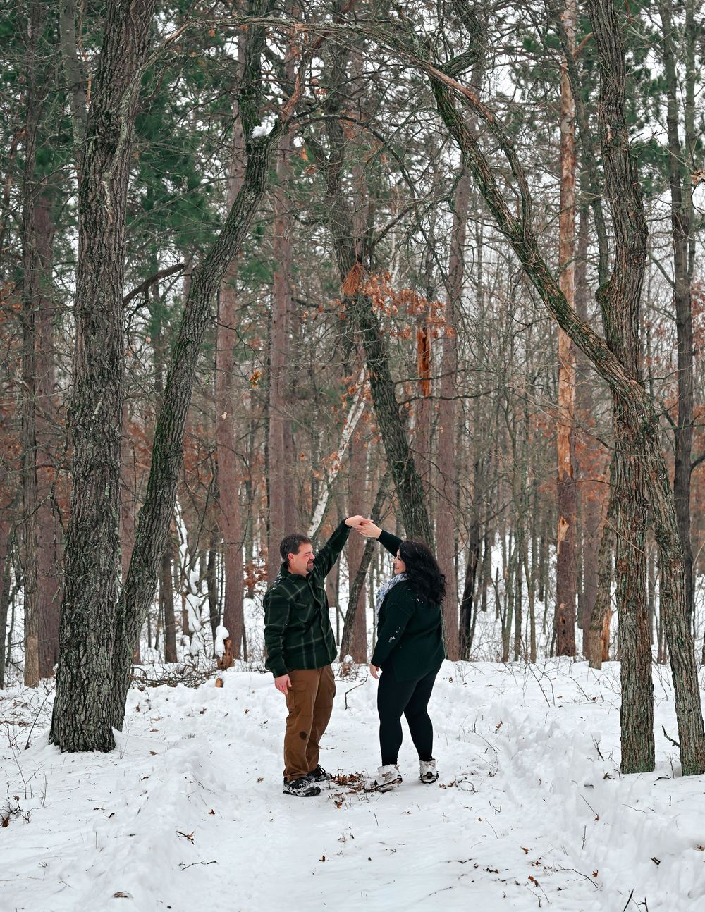 Couple dancing together on snowy forest path surrounded by tall winter trees.