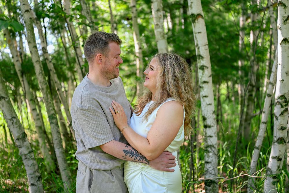 Couple embracing and smiling at each other in a birch tree forest, man in gray shirt, woman in white dress.