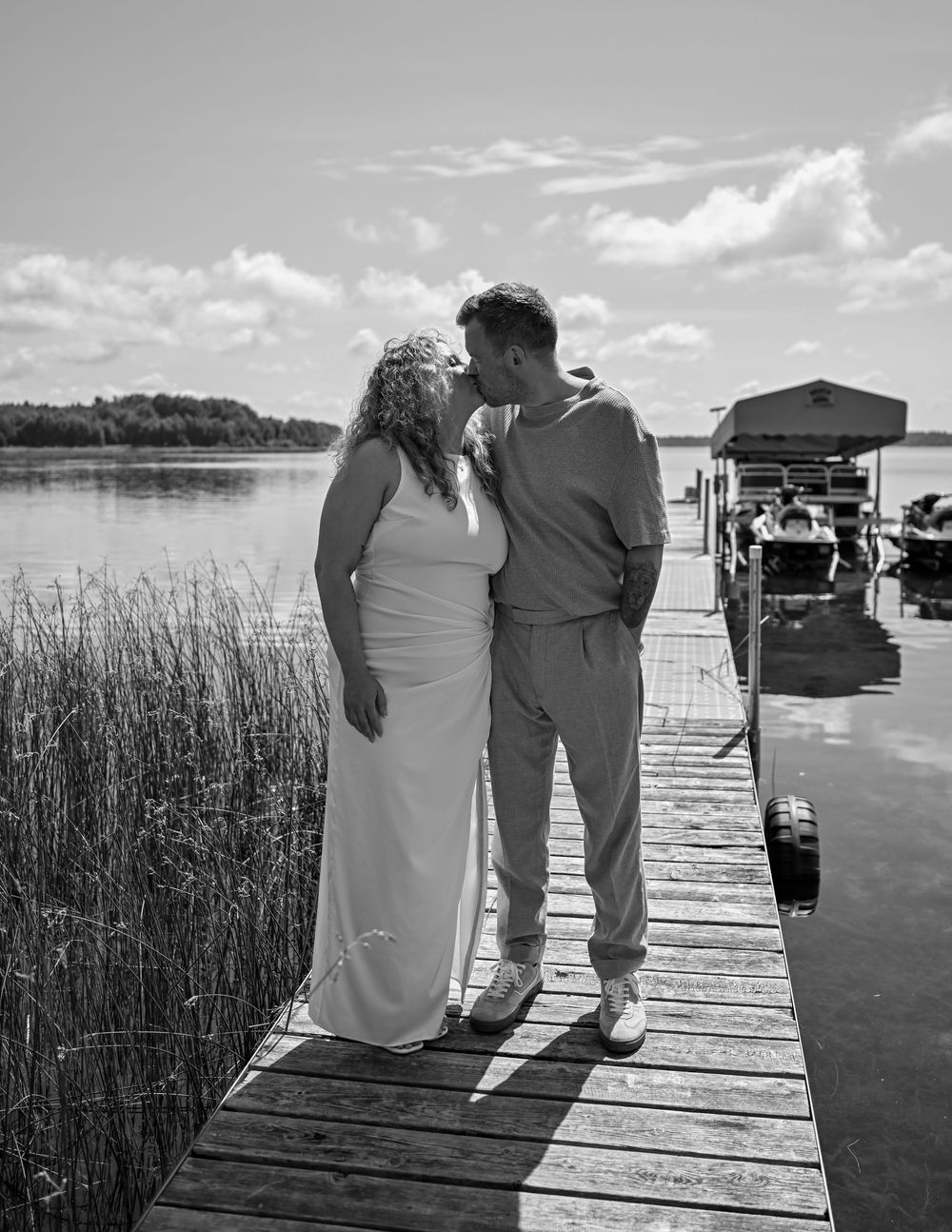 Black and white photo of a couple kissing on a lakeside dock, bride in white gown, groom in suit, on a sunny day.