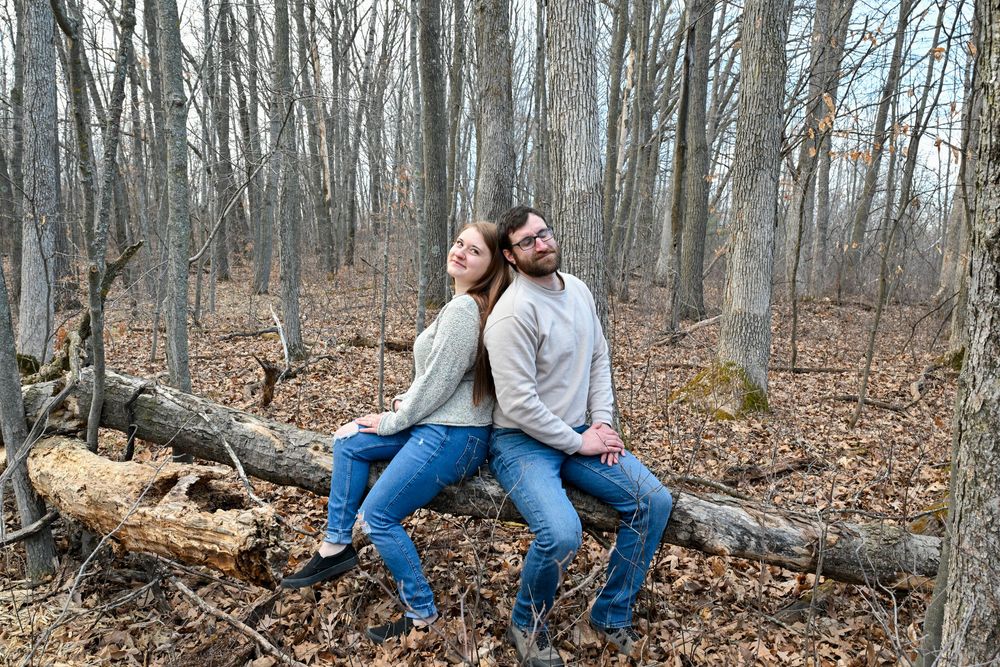 Happy couple sitting back-to-back on fallen log in leafless autumn forest smiling.