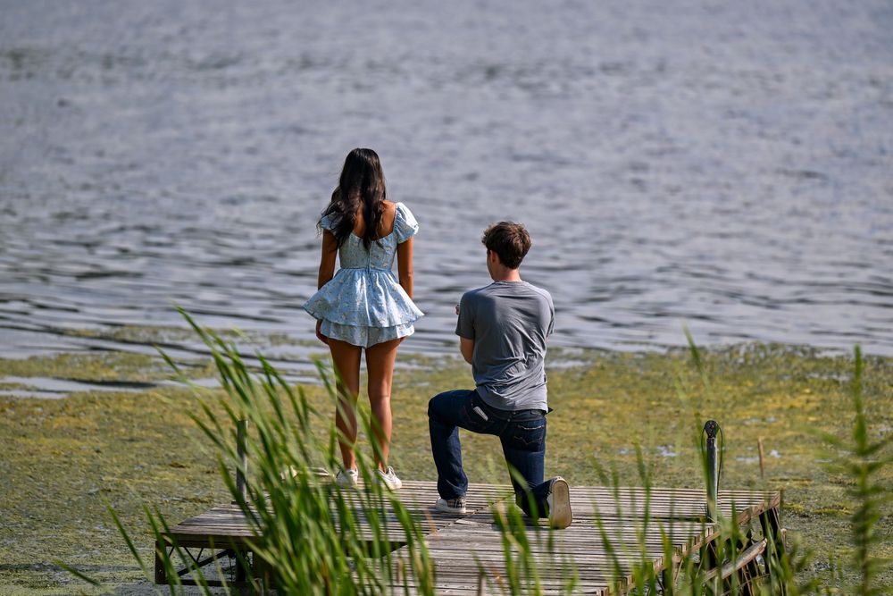 Man kneeling on a lakeside dock proposing to a woman in a blue floral dress near the water.