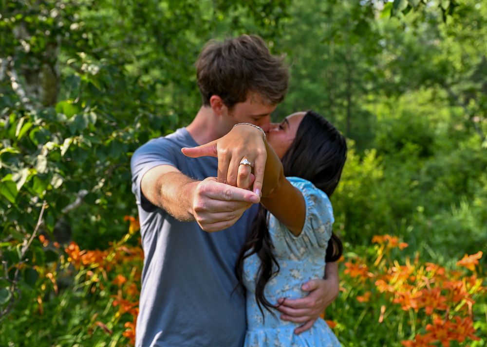Couple kissing while showing off an engagement ring surrounded by orange flowers.
