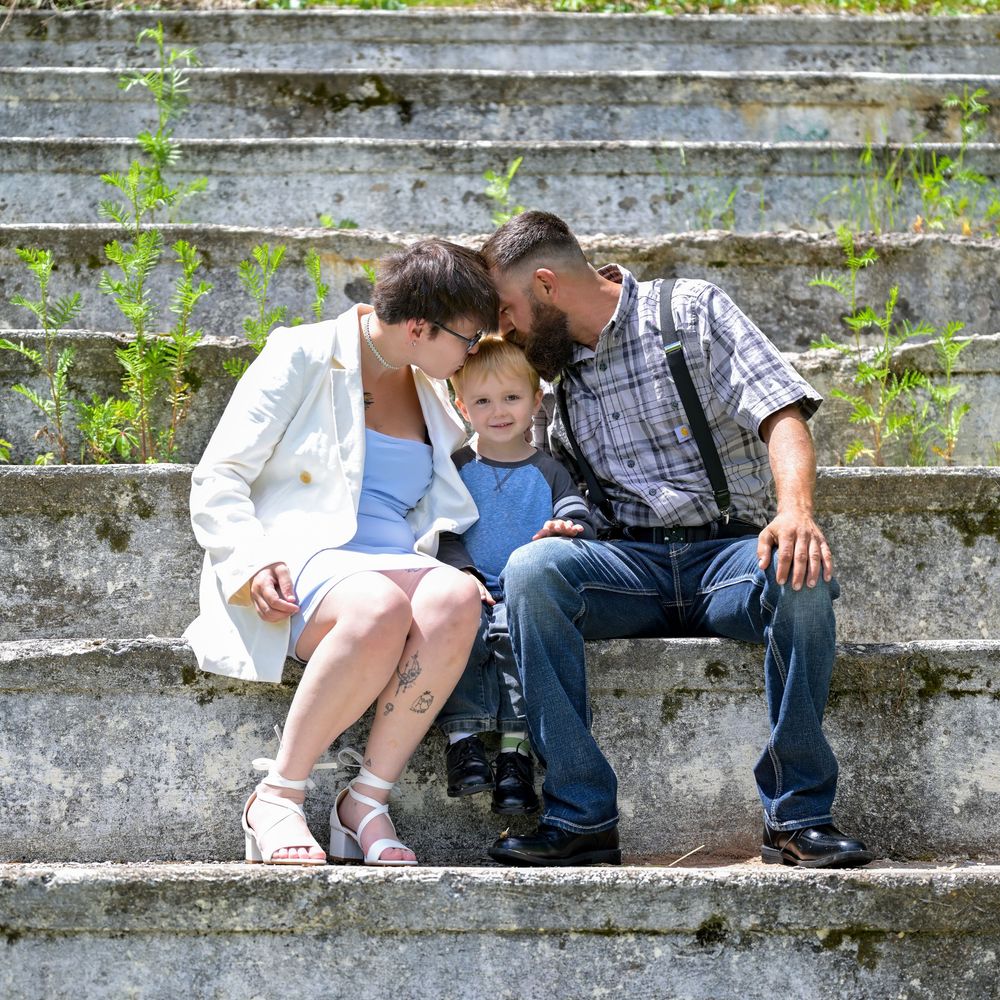 A family of three sits on stone steps outdoors, parents leaning in to embrace their young child in a sunny setting.