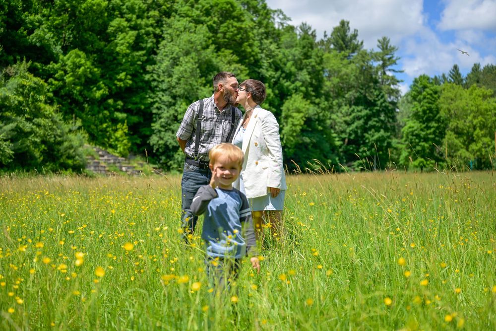 Family of three enjoying a sunny day in a lush green meadow with yellow wildflowers and trees in background.