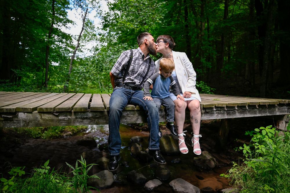 Family of three sitting on a wooden bridge in a lush green forest, parents kissing over their young child.