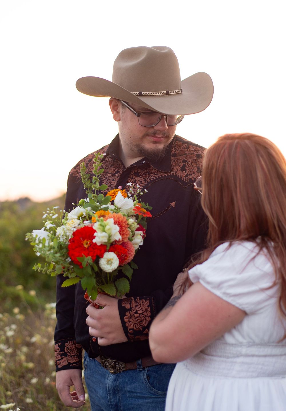 A couple shares an intimate moment in a field as the person in western attire holds a vibrant bouquet of flowers.