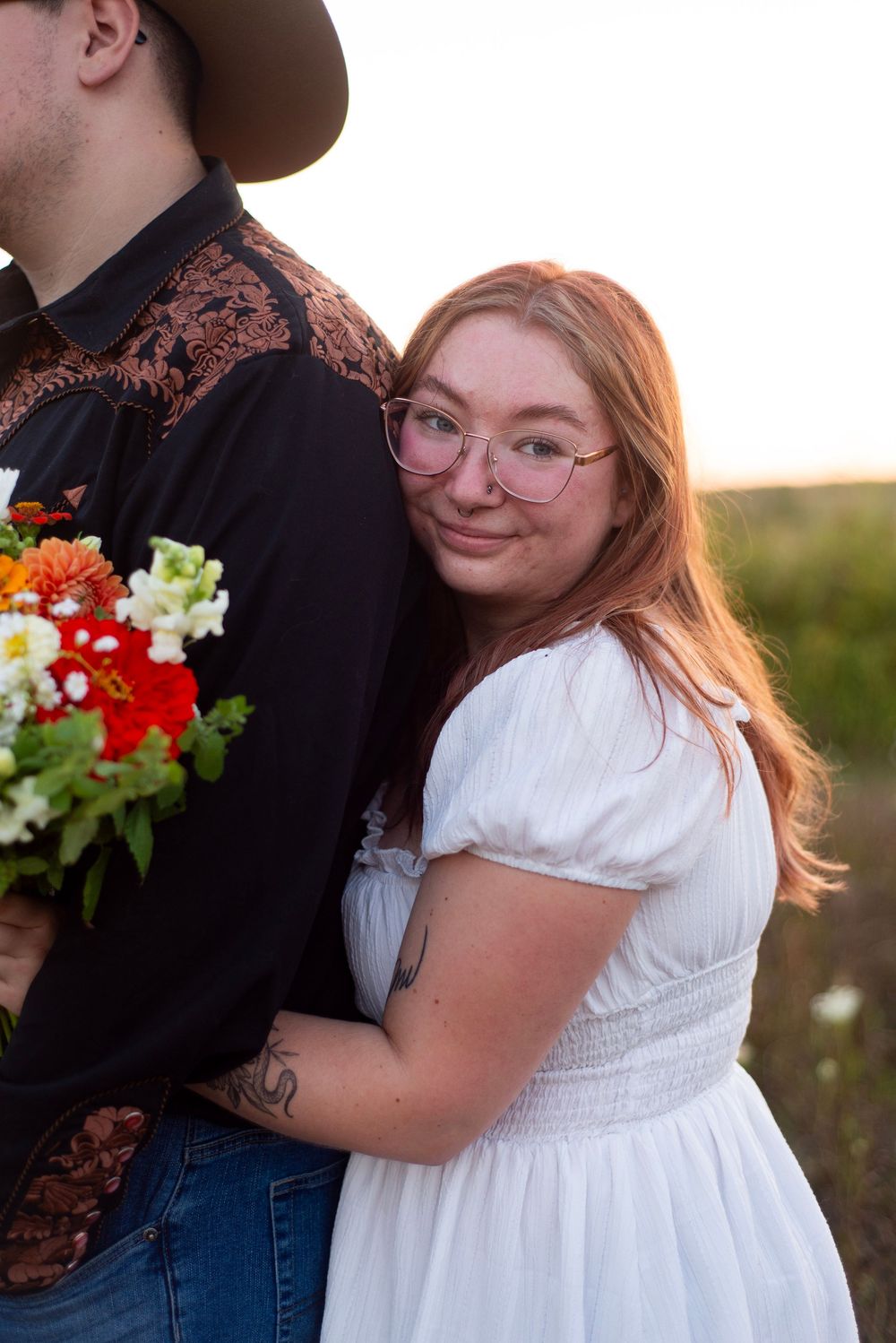 A couple in western wear embrace outdoors at sunset, with her wearing a white dress and him in dark clothing.