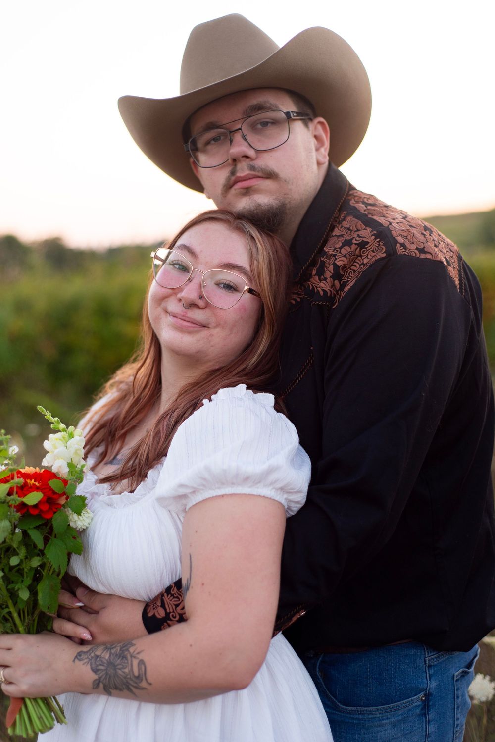 Couple shares intimate moments during rustic outdoor photoshoot with red floral bouquet at sunset.