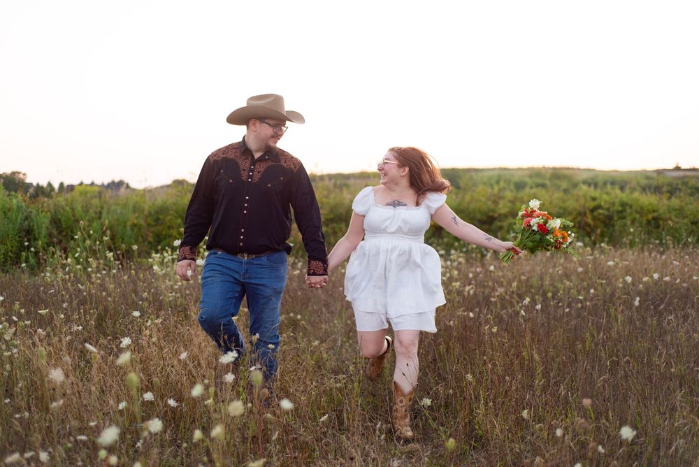 A couple in western and white attire walks hand in hand through a wildflower field together.