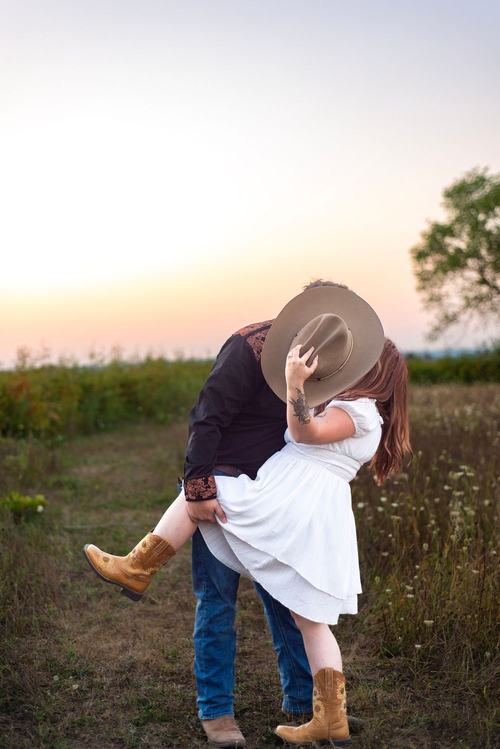 Playful couple in cowboy boots poses for romantic dip pose during sunset field photoshoot.