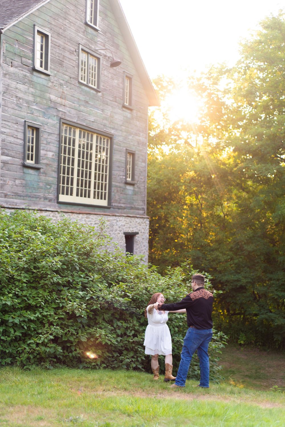Two people stand near a stone house at sunset as golden light filters through trees in the background.
