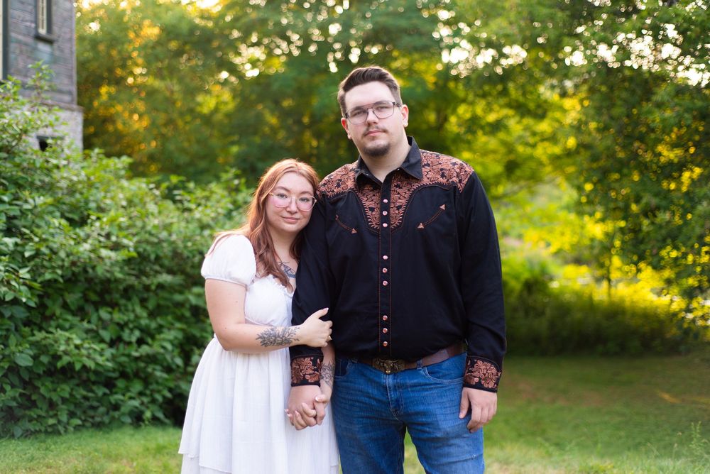 A couple poses together on a sunny day in a lush green outdoor setting wearing casual attire.