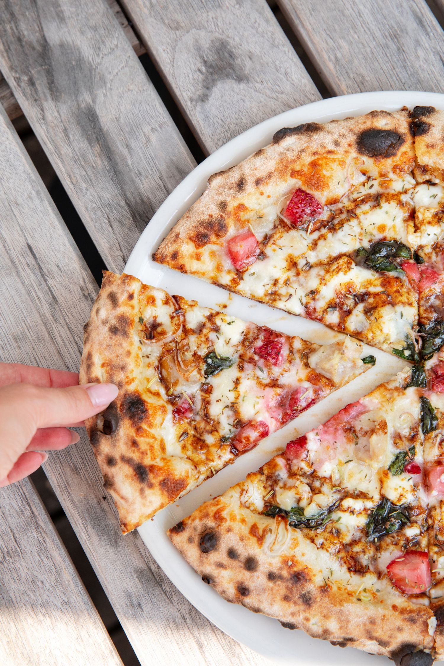 Wood-fired pizza with melted cheese, tomatoes, and fresh herbs being served on a wooden deck.