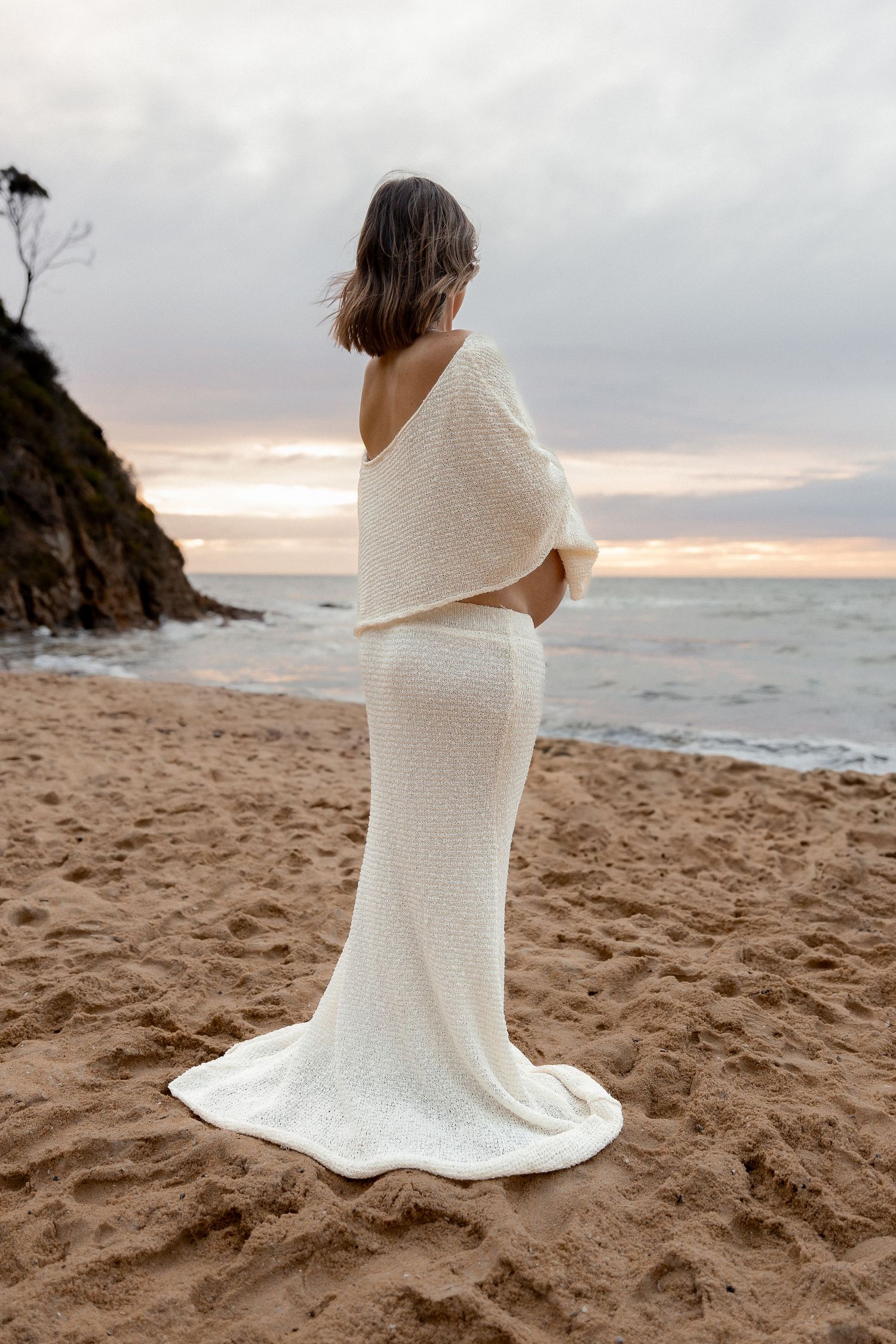 Person wrapped in white fabric stands alone on beach at sunset.