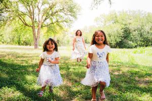 Three girls in white dresses running and laughing through a sunny green park with trees in the background.