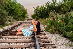 A woman in a striped dress and boots poses on railroad tracks surrounded by cacti and lush greenery.