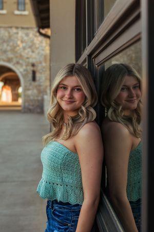Young blonde woman in teal crochet tube top and jeans leans against a mirrored wall, smiling outdoors.